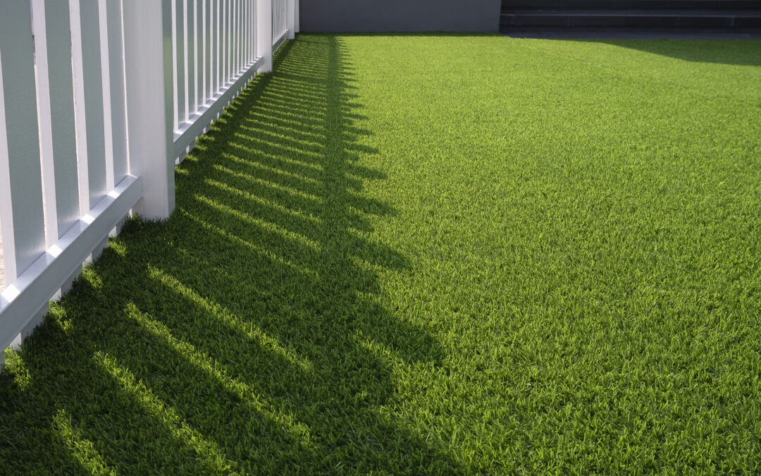 Sunlight and shadow on a white wooden fence and green artificial grass lawn in the front yard of a home for artificial-grass-in-Maryland blog,