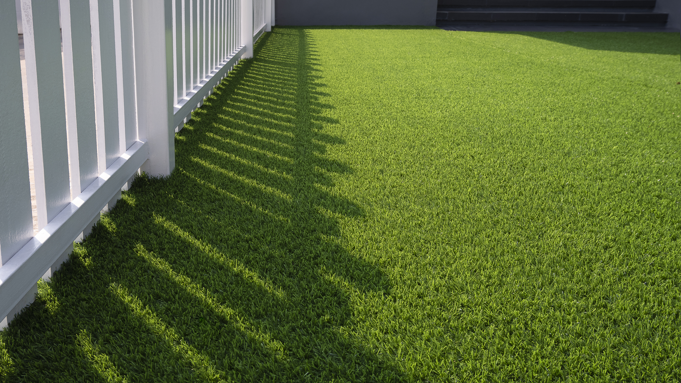 Sunlight and shadow on a white wooden fence and green artificial grass lawn in the front yard of a home for artificial-grass-in-Maryland blog,